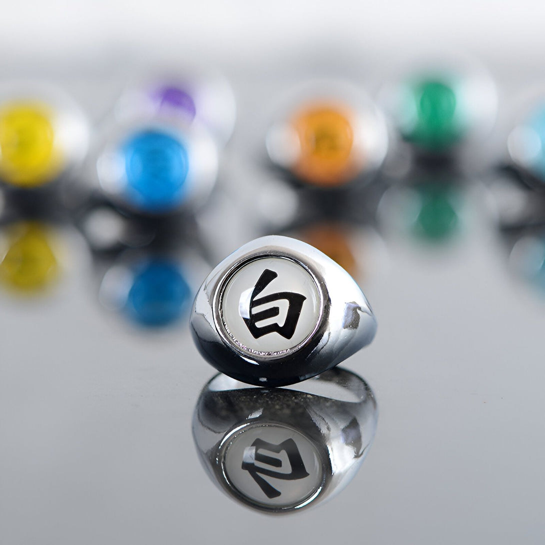 Bague en métal argenté avec le symbole "Byakugan" de l'anime Naruto, sur fond flou de plusieurs autres bagues avec des symboles différents.