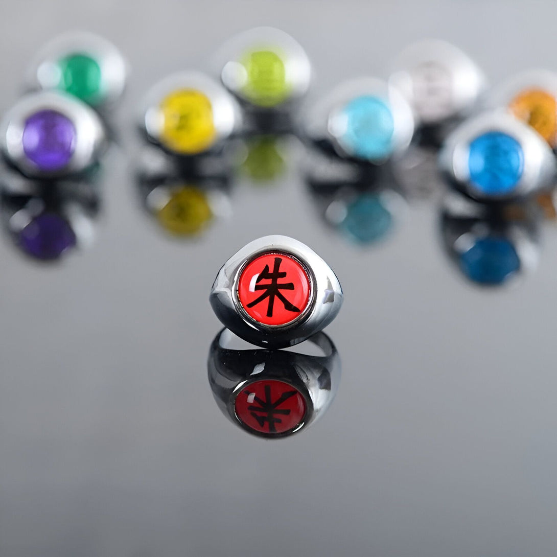 Bague de Sharingan rouge avec le symbole "Sharingan" en noir, sur un fond argenté. La bague est entourée d'autres bagues de Sharingan de différentes couleurs, reflétant dans un effet miroir.  Parfait pour les fans de Naruto !