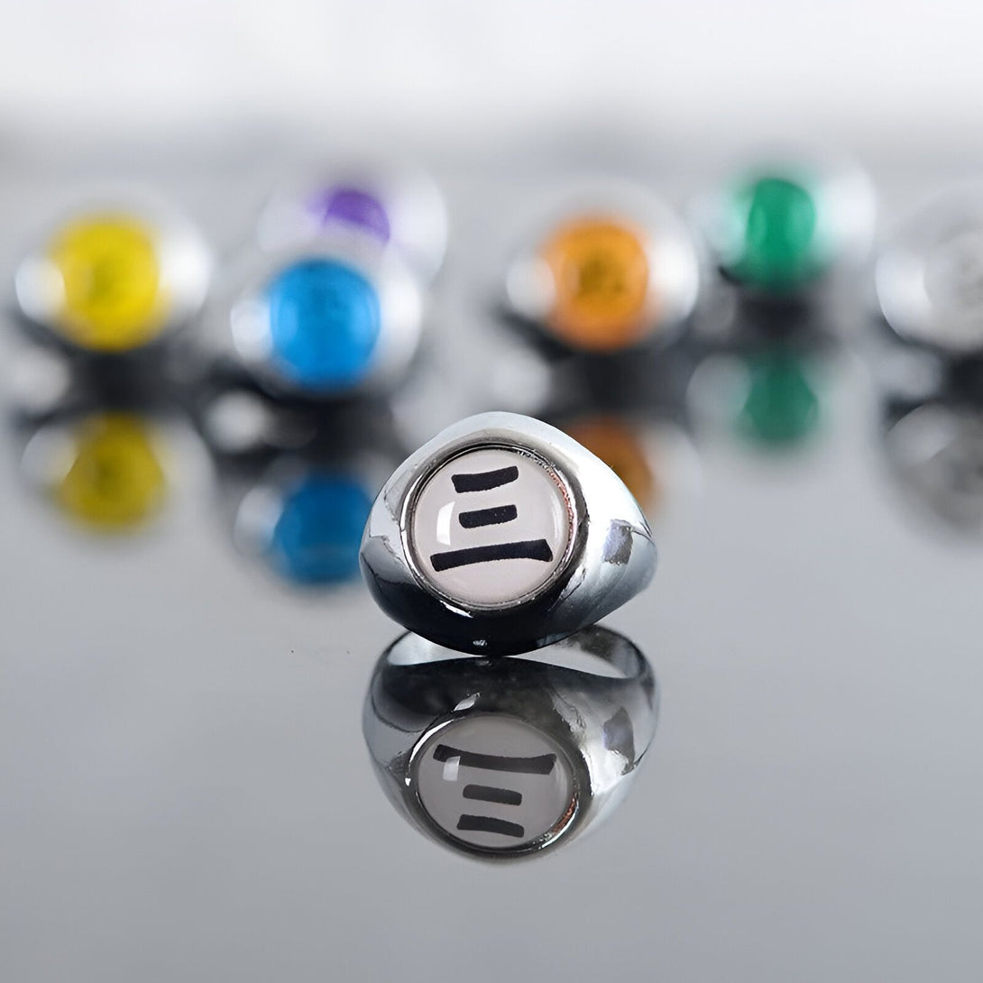 Bague en métal argenté avec le symbole du village caché de la feuille sur un fond réfléchissant. La bague est entourée d'autres bagues colorées.