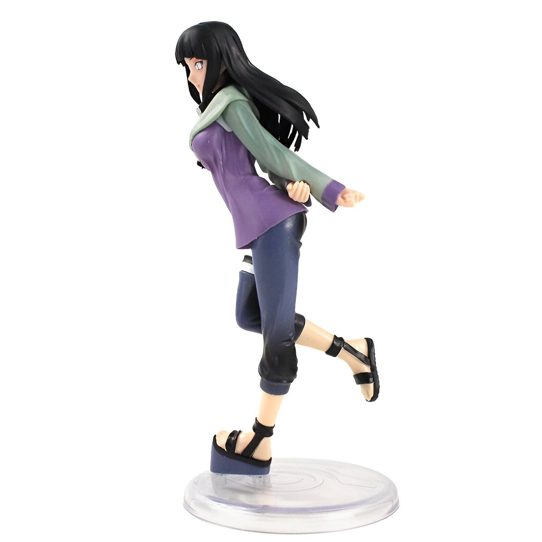 Figurine de Hinata Hyuga de l'anime Naruto, portant une tenue violette et verte, les cheveux noirs et les yeux violets. La figurine est en position dynamique, la jambe droite levée, le pied droit en l'air. Elle est posée sur un socle transparent avec le symbole du village caché de Konoha.