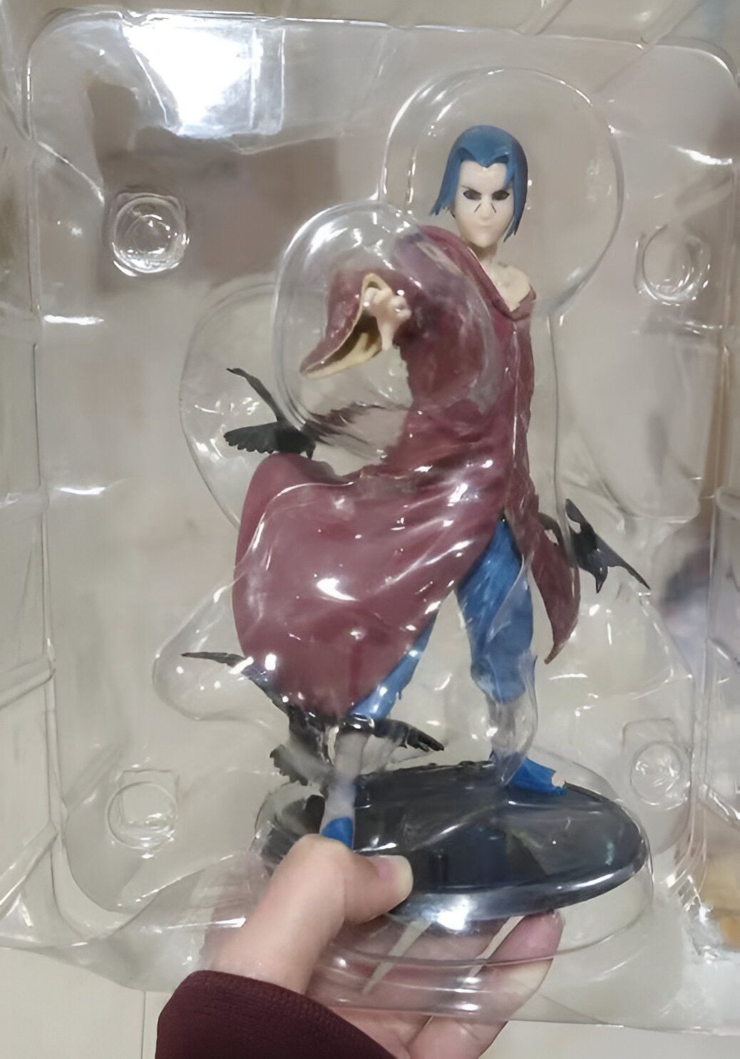 Figure de collection de Sasuke Uchiha, personnage du manga Naruto, dans son emballage d'origine. Sasuke est représenté en tenue de combat avec des corbeaux noirs à ses côtés.