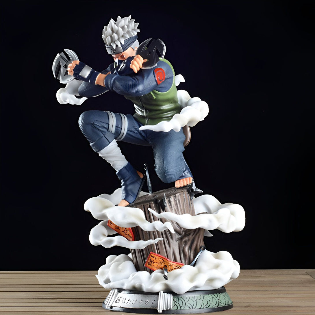 Figurine de Kakashi Hatake de Naruto, en position de combat, tenant des kunais et entouré de fumée blanche. La figurine est posée sur une base en bois avec des feuilles vertes.