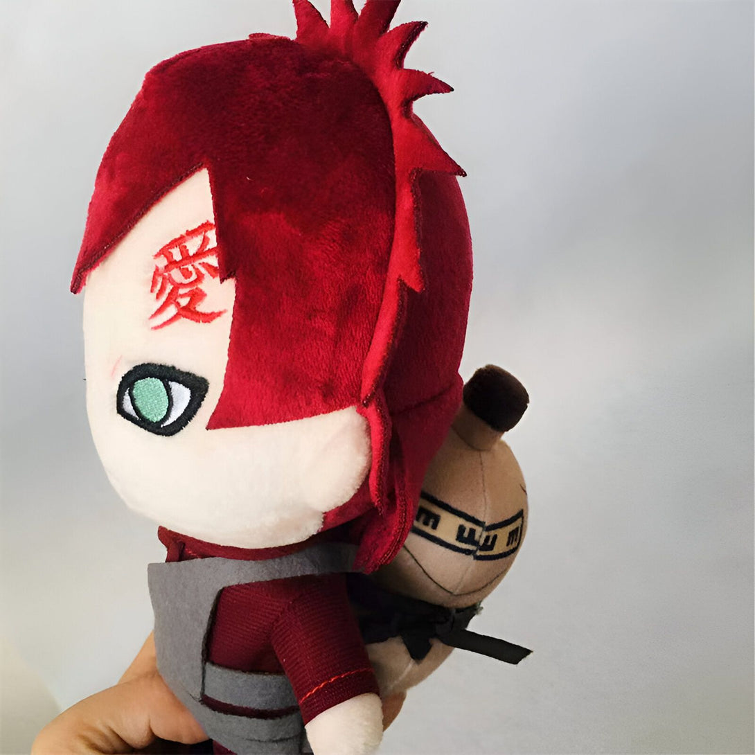 Peluche en forme de personnage de l'anime Naruto, portant une veste rouge et un pantalon gris avec un symbole rouge sur le front. Il porte également un petit sac à dos beige sur le dos.