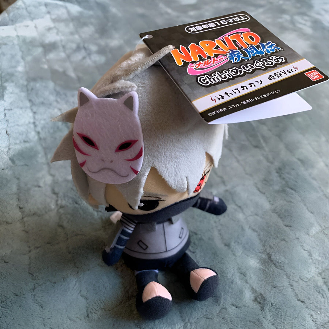 Peluche en forme de Kakashi Hatake de la série Naruto, portant un masque de  Sharingan rouge et blanc.  Il est assis et  tient une étiquette avec l'écriture "Naruto".  C'est un excellent produit pour les fans de Naruto.
