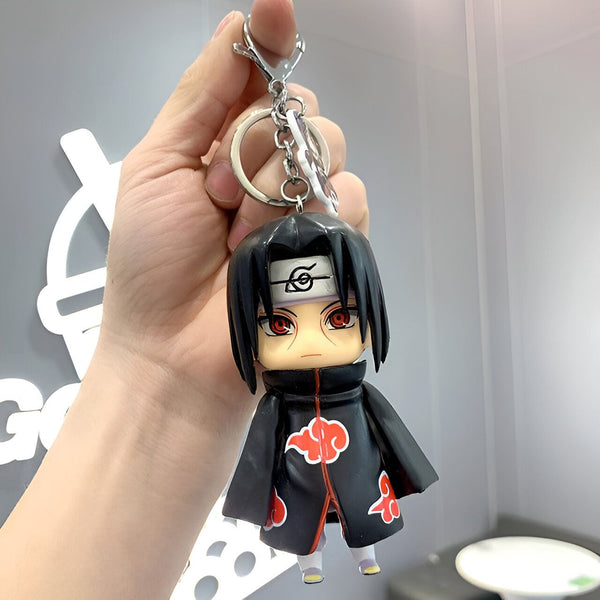 Un porte-clés en forme de figurine de Itachi Uchiha, un personnage de l'anime Naruto, portant son célèbre costume noir avec le symbole du clan Uchiha sur sa veste.  La figurine est attachée à un anneau et une chaîne en argent.