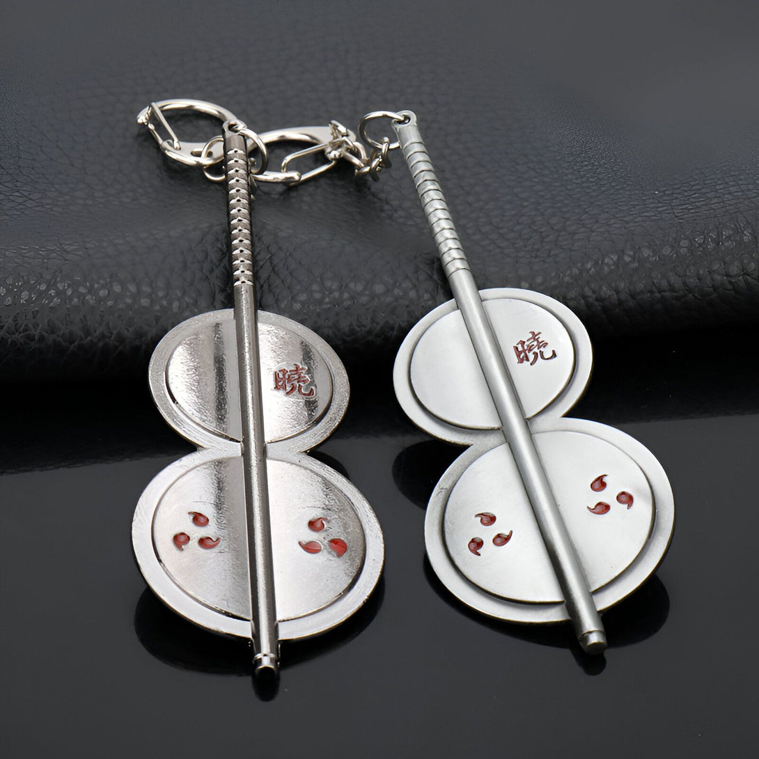 Porte-clés en métal argenté représentant le symbole du Sharingan de Naruto, un œil rouge avec trois tomoes noirs. Le porte-clés est composé de deux disques ronds reliés par une barre. Les disques sont décorés d'un kanji écrit en rouge.