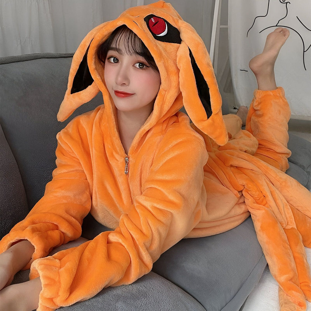 Une jeune femme portant une combinaison en peluche orange avec une capuche représentant le démon renard à neuf queues de Naruto. La combinaison est zippée sur le devant et présente des détails en fourrure. La femme est assise sur un canapé gris.