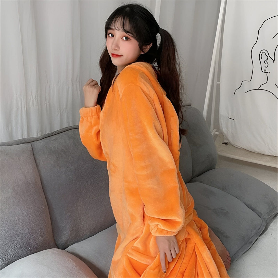 Une jeune femme souriante porte une robe de molleton orange à capuche. Elle est assise sur un canapé gris clair dans une pièce lumineuse.