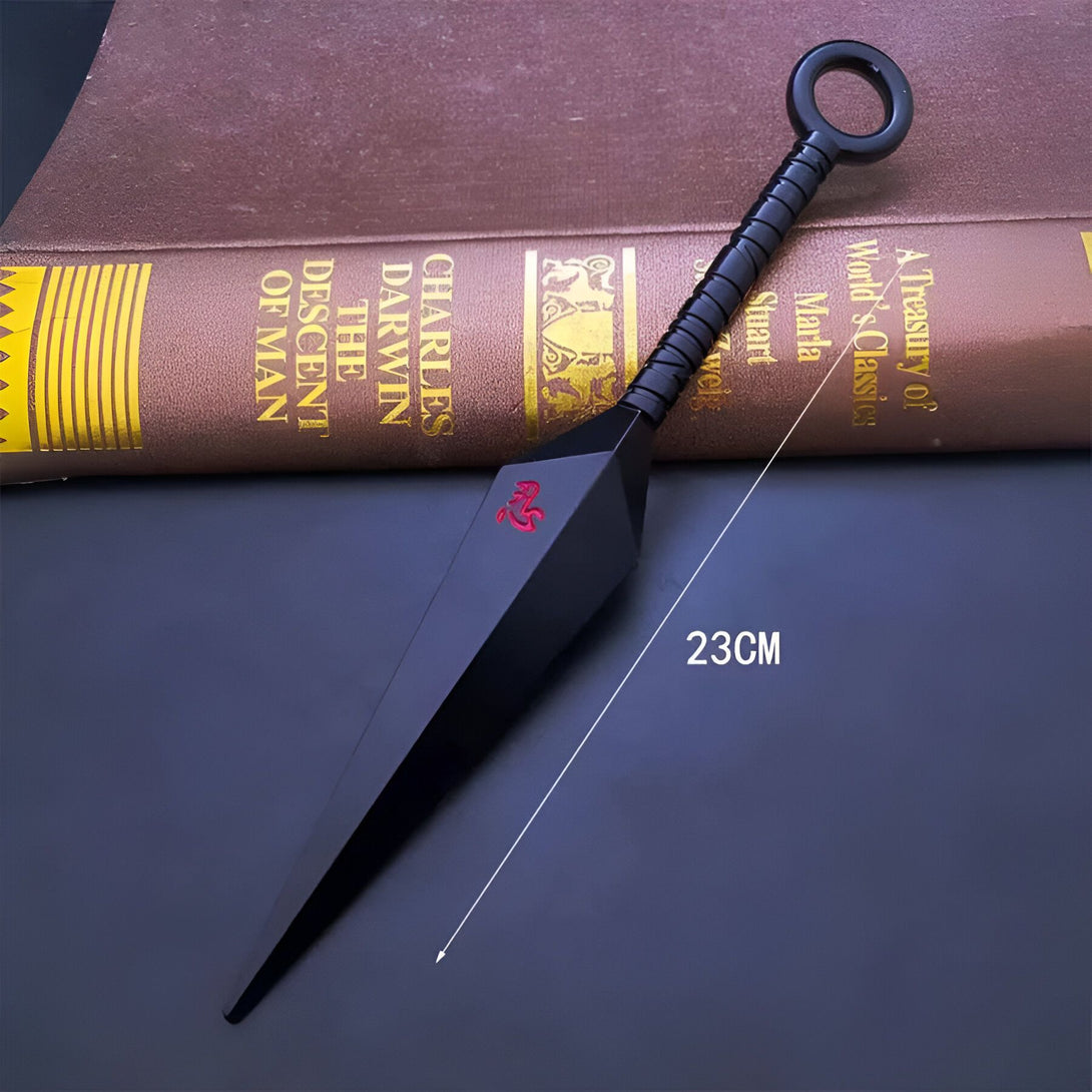 Une  kunai noire  de 23 cm avec un anneau  sur la  poignée.  La kunai est  posée sur un livre.  La kunai est  une arme de lancer  utilisée  dans la série manga et anime  Naruto.  C'est un objet  populaire pour  les fans de Naruto.