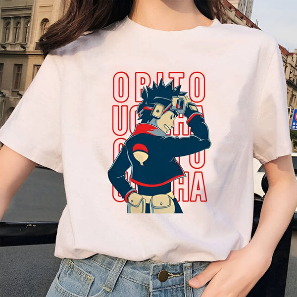 Une femme porte un t-shirt blanc avec un imprimé du personnage Obito Uchiha de l'anime Naruto. Le t-shirt est à manches courtes et présente un imprimé coloré d'Obito dans son uniforme d'Akatsuki. Le nom "Obito Uchiha" est écrit en rouge sur le t-shirt.